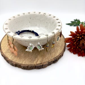 May include: A collection of earrings displayed on a white ceramic bowl with a speckled finish. The earrings include various designs, such as daisy-shaped, beaded, and feather-like styles. The bowl sits on a wooden slice, with a dark blue bracelet inside.