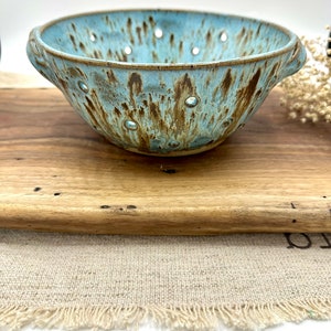 May include: A handmade ceramic colander with a light blue glaze and brown accents. The bowl has a series of drainage holes and two handles. The colander is sitting on a wooden cutting board.