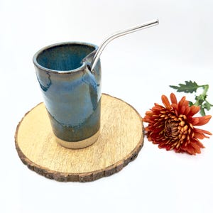 Peut inclure: Une tasse en céramique bleue et brune avec une paille en acier inoxydable. La tasse est posée sur un dessous de verre en bois. Une seule fleur orange est en arrière-plan.