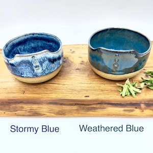 May include: Two handmade ceramic bowls in shades of blue, labeled "Stormy Blue" and "Weathered Blue." Each bowl has a unique shape with a cutout on one side and three small decorative holes. The bowls sit on a wooden surface.