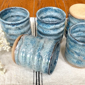 May include: Five blue and white ceramic mugs with a wavy design. The mugs are arranged in a semi-circle on a white and black striped surface.