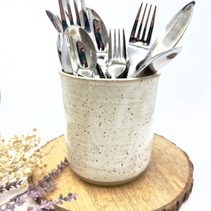 Op de afbeelding: Een keramische keukengerei houder met een gespikkelde, gebroken witte afwerking, gevuld met glanzend zilverwerk. De houder staat op een ronde houten onderzetter. Gedroogde bloemen en lavendel takjes zijn in de buurt geplaatst.