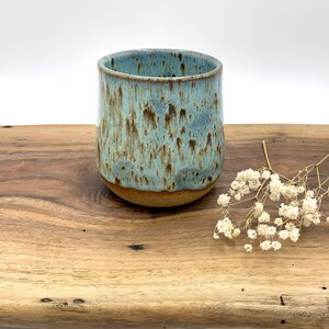 May include: A handmade ceramic tumbler with a speckled blue and brown glaze. The cylindrical cup has a rounded base and is displayed on a wooden surface. A sprig of white flowers is placed to the side.