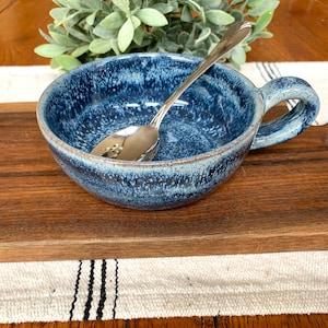 Può includere: Una ciotola di zuppa in ceramica blu con una glassa macchiettata e un manico. La ciotola è appoggiata su un tagliere di legno con un cucchiaio all'interno.