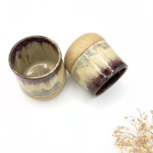 May include: Two ceramic mugs with a brown, beige, and white glaze. The mugs have a rounded shape and a slightly flared lip.