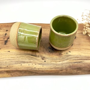 以下が含まれることがあります： 緑色の釉薬と素焼きのベースが特徴の手作りのセラミックカップが2つ。1つは直立し、もう1つは横になっています。カップは木製の表面に置かれ、陶器の土の色合いと質感を際立たせています。