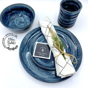 May include: A set of blue stoneware dinnerware with a white napkin and a sprig of greenery. The dinnerware is handmade by Tessa Rohde and features a speckled blue glaze. The napkin is folded and tied with twine. The text "Made in USA" and "Colonial Collections Stoneware" is visible on a small tag.