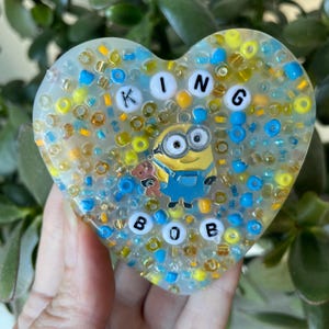 Op de afbeelding: Een hartvormige harsversiering met een geel en blauw Minion-personage en de woorden "KING BOB" gespeld in witte letters. De achtergrond is gevuld met kleine, kleurrijke kralen.
