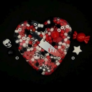 May include: A heart-shaped resin tray filled with red, white, black, and silver beads. A small knife with a blood splatter design is embedded in the resin. Additional decorative elements include a skull, a star, and a sweet.