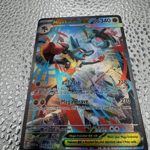 May include: A Mega Lucario EX trading card with 340 HP, featuring a dynamic illustration of the blue and black character in a fighting stance. The card displays the attacks Aura Jab and Mega Brave, with the set number 179/132 and a holographic finish.