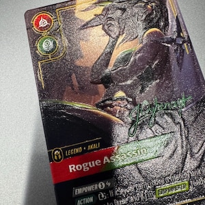 Può includere: Una carta collezionabile rettangolare con un'illustrazione scura e testurizzata di un personaggio. La carta mostra il testo Legend Akali, Rogue Assassin ed Empower 3. Include icone circolari rosse e verdi, una firma e testo di gioco.