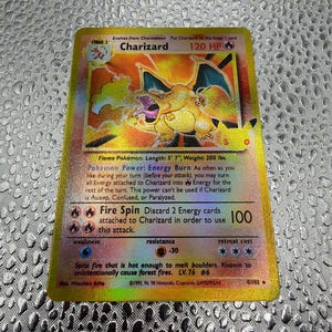 May include: A gold-bordered Charizard trading card featuring the fire-breathing dragon illustration, 120 HP, Energy Burn power, and Fire Spin attack. The card displays the 25th-anniversary logo, copyright text, and 4/102 numbering.