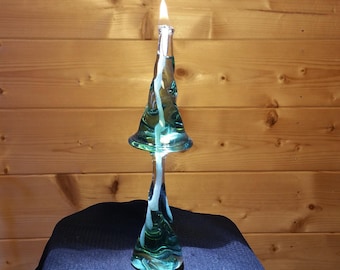 Könnte beinhalten: Eine tealfarbene Glas-Öllampe mit einem brennenden weißen Docht. Die Lampe ist in Form eines Pilzes mit einem breiten, ausgestellten Oberteil und einem schlanken Stiel geformt. Die Lampe steht auf einer schwarzen Stoffoberfläche.