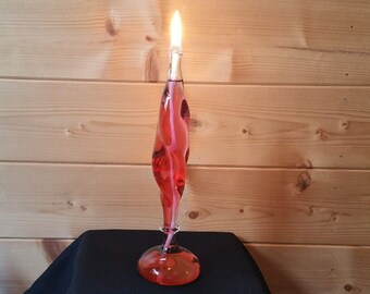 Könnte beinhalten: Eine rote Glas-Öllampe mit einem brennenden weißen Docht. Die Lampe steht auf einer schwarzen Stoffoberfläche.