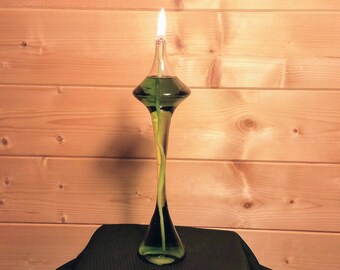 Könnte beinhalten: Eine hohe, grüne Glasöllampe mit geschwungener Form und brennendem Docht. Die Lampe enthält grüne Flüssigkeit und steht auf einer schwarzen, mit Stoff bedeckten Oberfläche vor einem Holz Hintergrund.