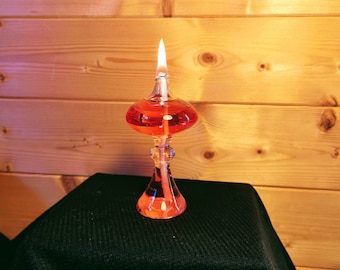 Könnte beinhalten: Eine rote Glas-Öllampe mit einem klaren Glasfuß und einem Docht, der mit einer hellen Flamme brennt. Die Lampe steht auf einer schwarzen Oberfläche.