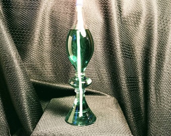 Könnte beinhalten: Eine tealfarbene Glas-Öllampe mit einem weißen Docht, der brennt. Die Lampe steht auf einer schwarzen, strukturierten Oberfläche.