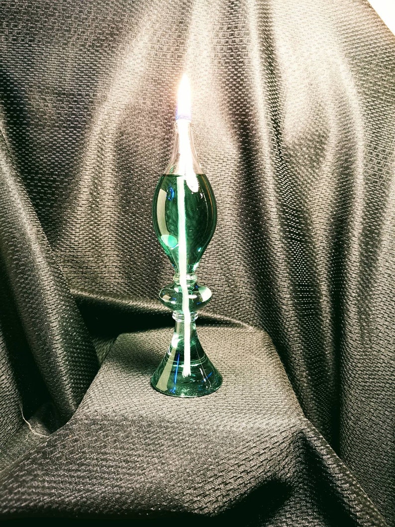 May include: A teal glass oil lamp with a white wick burning. The lamp is on a black textured surface.