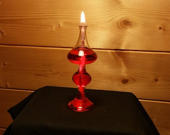 Könnte beinhalten: Eine rote Glas-Öllampe mit einem weißen Docht, der brennt. Die Lampe steht auf einer schwarzen Oberfläche.