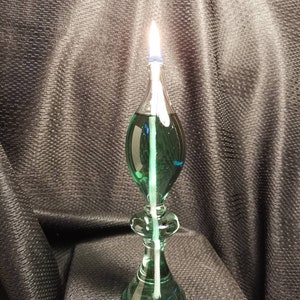 May include: A green glass oil lamp with a white wick and a blue glass top. The lamp is lit and the flame is visible.