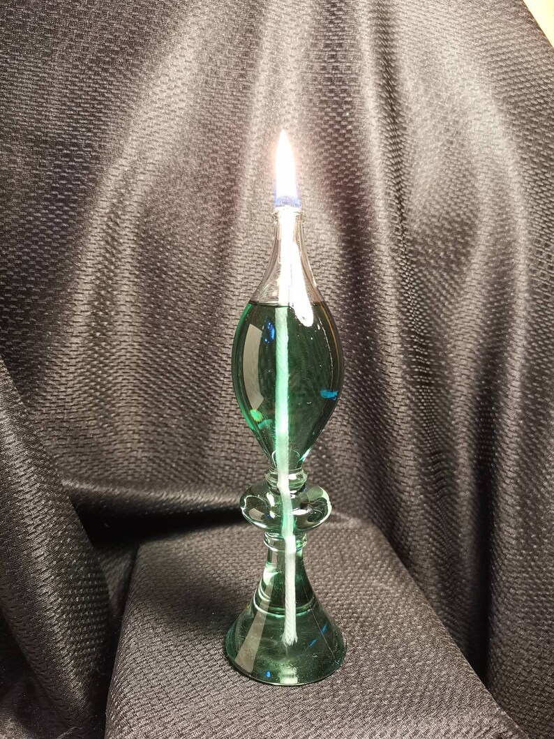 May include: A green glass oil lamp with a white wick and a blue glass top. The lamp is lit and the flame is visible.