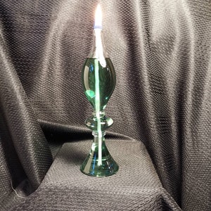 May include: A green glass oil lamp with a white wick burning. The lamp has a round base and a teardrop-shaped body.