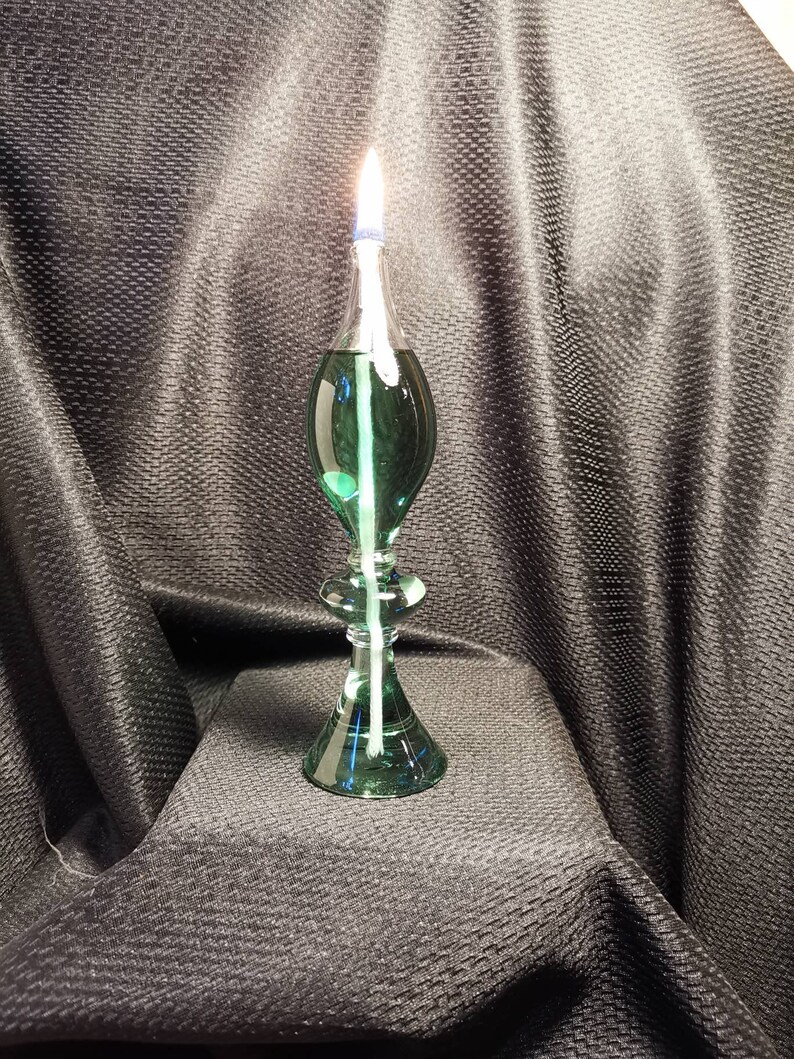May include: A green glass oil lamp with a white wick burning. The lamp has a round base and a teardrop-shaped body.