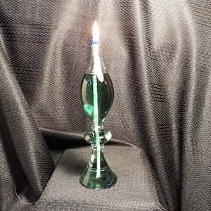 May include: A green glass oil lamp with a white wick and a blue glass base. The lamp is lit and the flame is visible.