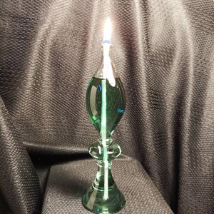 May include: A green glass oil lamp with a white wick burning. The lamp has a round base and a teardrop-shaped body.