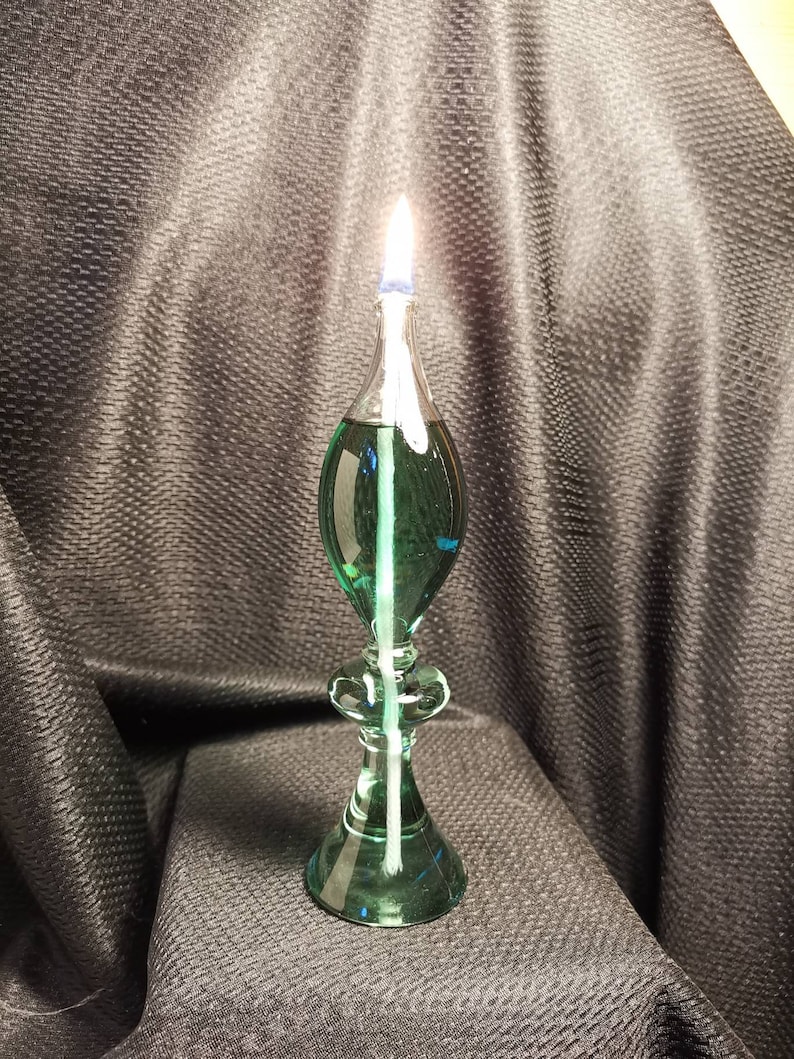 May include: A green glass oil lamp with a white wick burning. The lamp has a round base and a teardrop-shaped body.