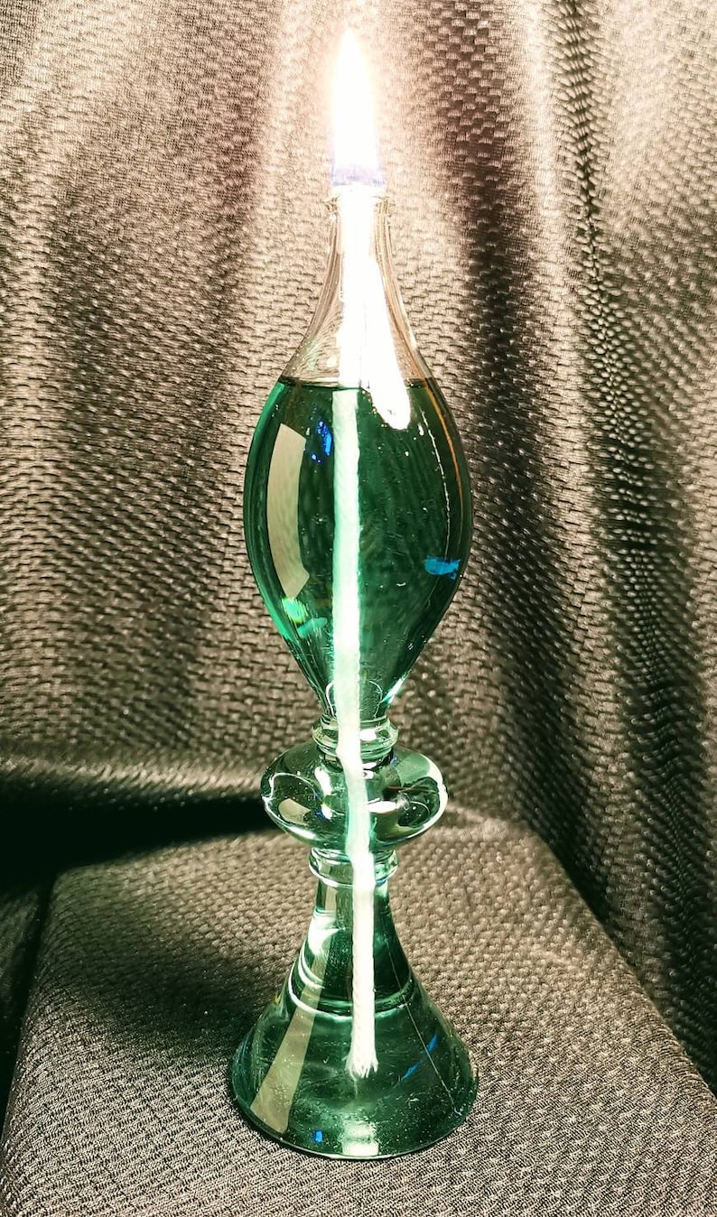 May include: A green glass oil lamp with a white wick and a clear glass reservoir. The lamp is lit and the flame is visible.