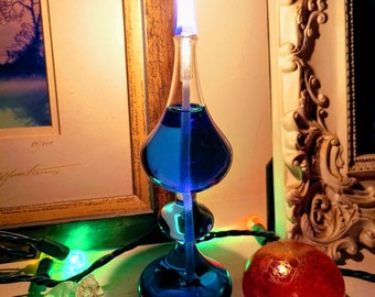 Könnte beinhalten: Eine blaue Glas-Öllampe mit einer Flamme, die oben brennt. Die Lampe steht auf einem Tisch mit einer roten Kugel und einem durchsichtigen Glasornament.