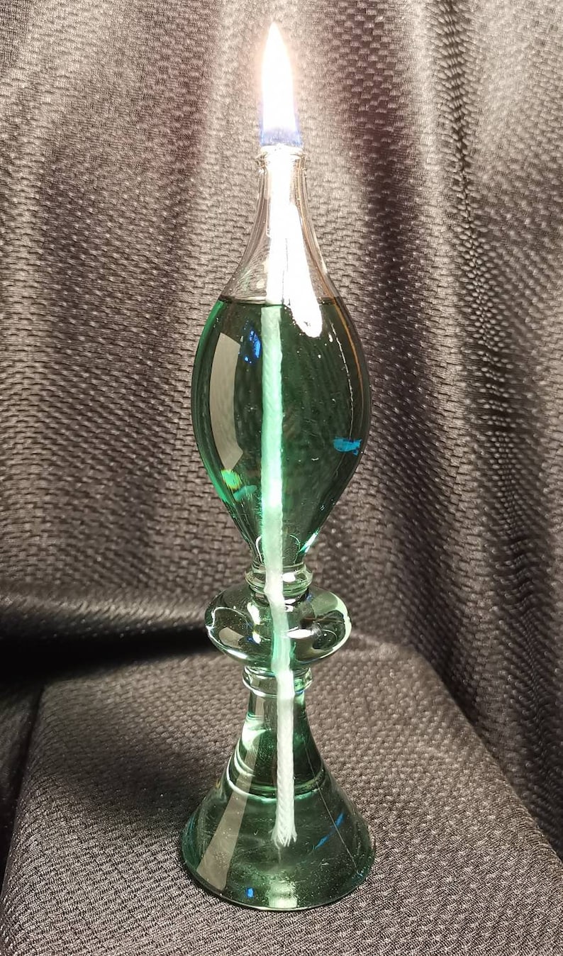 May include: A green glass oil lamp with a white wick and a clear glass reservoir. The lamp is lit and the flame is visible.