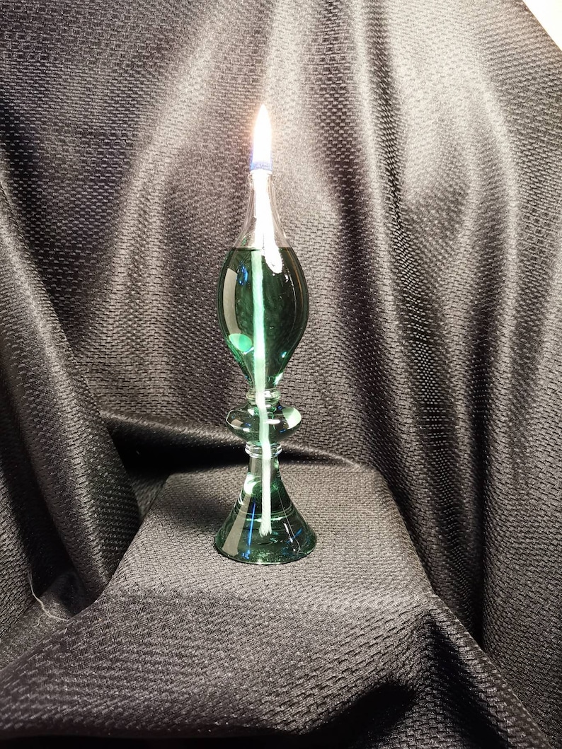 May include: A green glass oil lamp with a white wick burning. The lamp has a round base and a teardrop-shaped body.