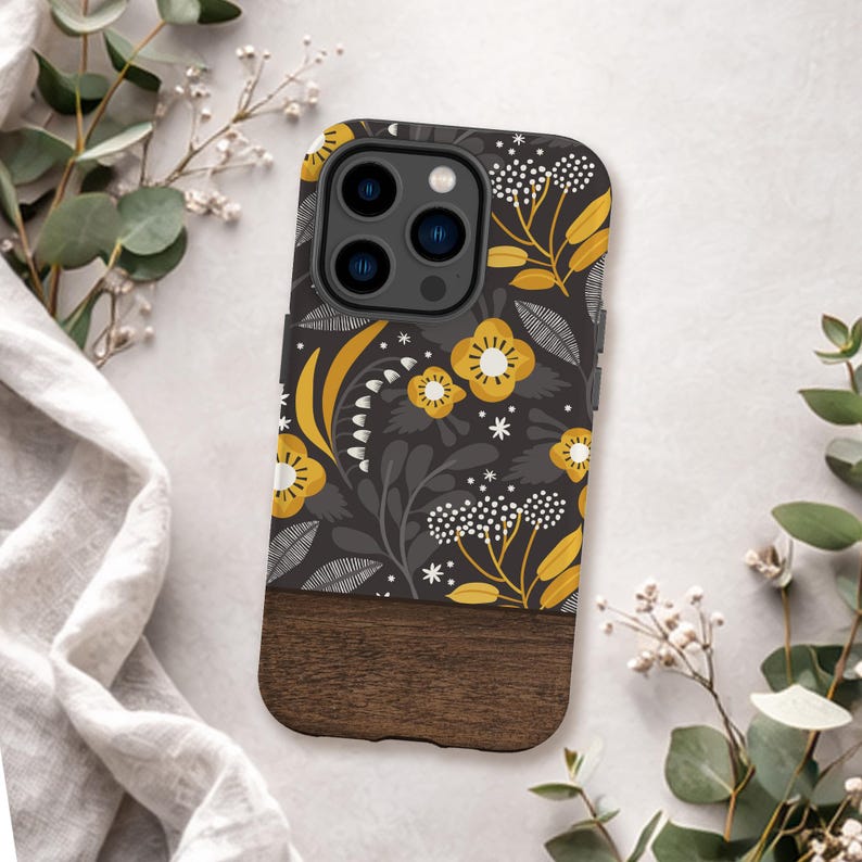May include: A phone case featuring a dark gray background with a repeating pattern of yellow flowers, white floral clusters, and gray leaves. The bottom section of the case is made of dark brown wood, contrasting with the floral upper portion.