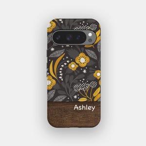 May include: A phone case featuring a dark gray background with a mustard yellow and white floral pattern. The bottom section has a dark brown wood grain texture with the name Ashley printed in white text across the center of the wood panel.