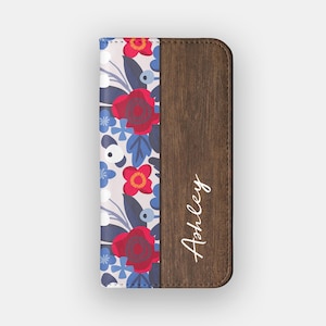 May include: A floral phone case with a wood grain finish. The left side features a pattern of red roses, blue flowers, and dark blue leaves on a light pink background. The right side has a brown wood grain finish with the name "Ashley" in white script.