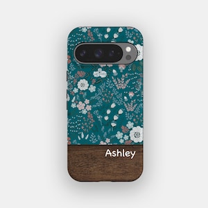 May include: A phone case featuring a dark teal background with a repeating pattern of white, light pink, and grey flowers and foliage. The bottom section consists of a dark brown wood grain texture with the name Ashley printed in white text.