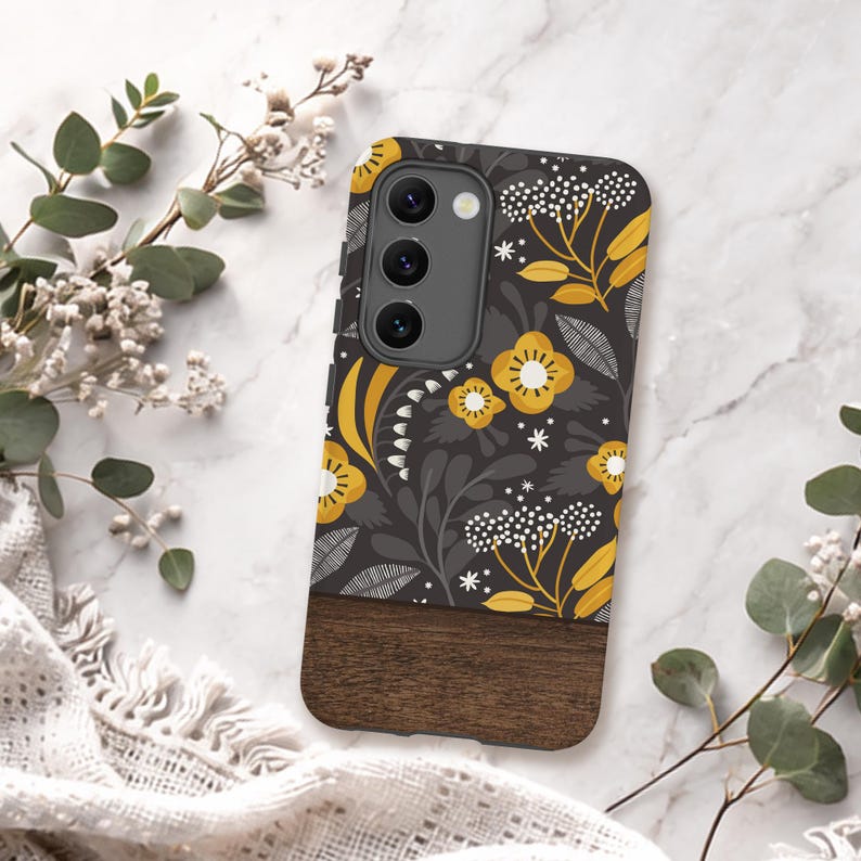 May include: A rectangular phone case featuring a dark gray background with a mustard yellow and white floral pattern on the top portion and a solid dark brown wood grain texture on the bottom portion, including a triple camera cutout.