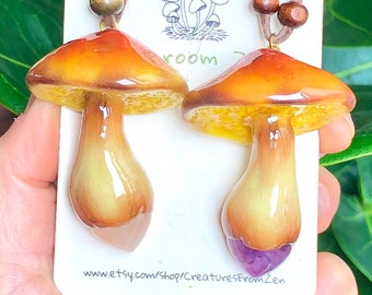 May include: Two ceramic mushroom necklaces with brown leather cords and wooden beads. One mushroom has a purple amethyst stone at the bottom of the stem. The necklaces are displayed on a white card with the text "www.etsy.com/shop/CreaturesFrom2en".