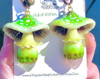May include: A pair of green and white mushroom earrings with a pink crystal charm. The earrings are on a brown cord necklace. The text "Shroom" is visible on the card.