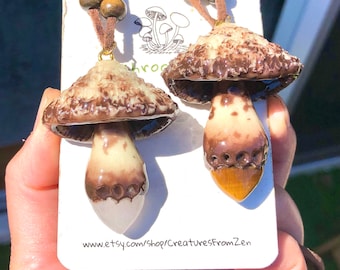 May include: Two brown and white mushroom necklaces with brown leather cords. The mushrooms have a textured surface and a brown cap with white spots. The necklaces are on a white card with the text "www.etsy.com/shop/CreaturesFromZen".