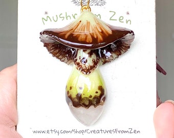 May include: A brown and white mushroom pendant necklace with a gold chain. The mushroom has a brown cap with a white stem and brown spots. The pendant is on a white card with the text "Mushroom Zen" and a website address.