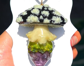 May include: A mushroom pendant with a black and white spotted cap, a yellow stem, and a green and purple base. The pendant hangs from a brown cord. The mushroom is on a white card.