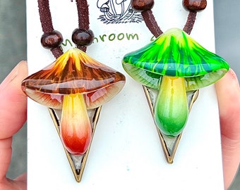 May include: Two mushroom-shaped necklaces with brown leather cords. One necklace features a green mushroom with white stripes and the other features a brown mushroom with a yellow and orange stem. The necklaces are on a white background with the text "www.etsy.com/shop/CreaturesFromZen".