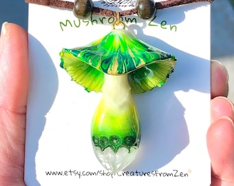 May include: A green and white mushroom pendant necklace with a brown leather cord and wooden beads. The pendant is made of resin and has a marbled effect. The necklace is labeled "Mushroom Zen" and includes the website "www.etsy.com/shop/creaturesfromzen".