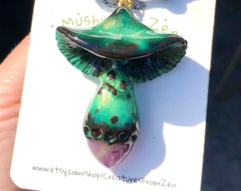 May include: A green and black mushroom pendant necklace with a purple amethyst stone at the bottom. The necklace is made of polymer clay and is attached to a brown leather cord. The pendant is about 2 inches tall. The text "Mush Zen" is written on the card behind the necklace. The website address "www.etsy.com/shop/CreaturesfromZen" is also written on the card.