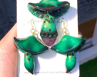 May include: A set of green and gold mushroom earrings and a pendant necklace. The earrings and pendant are made of polymer clay and have a shiny, iridescent finish. The pendant features a purple amethyst stone.
