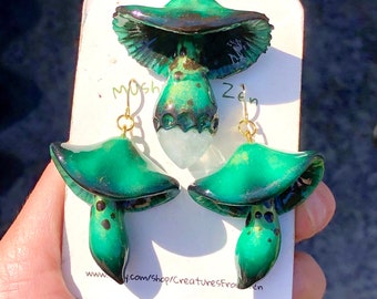 May include: A set of green mushroom earrings and a pendant necklace. The mushroom caps are a dark green with black accents and the stems are a lighter green with black spots. The pendant is attached to a gold chain.