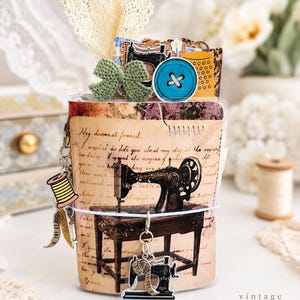 May include: A small journal featuring a vintage sewing machine illustration on the cover, accented with cursive text, a green crochet bow, a blue button, and dangling metal charms shaped like a spool of thread and a sewing machine.